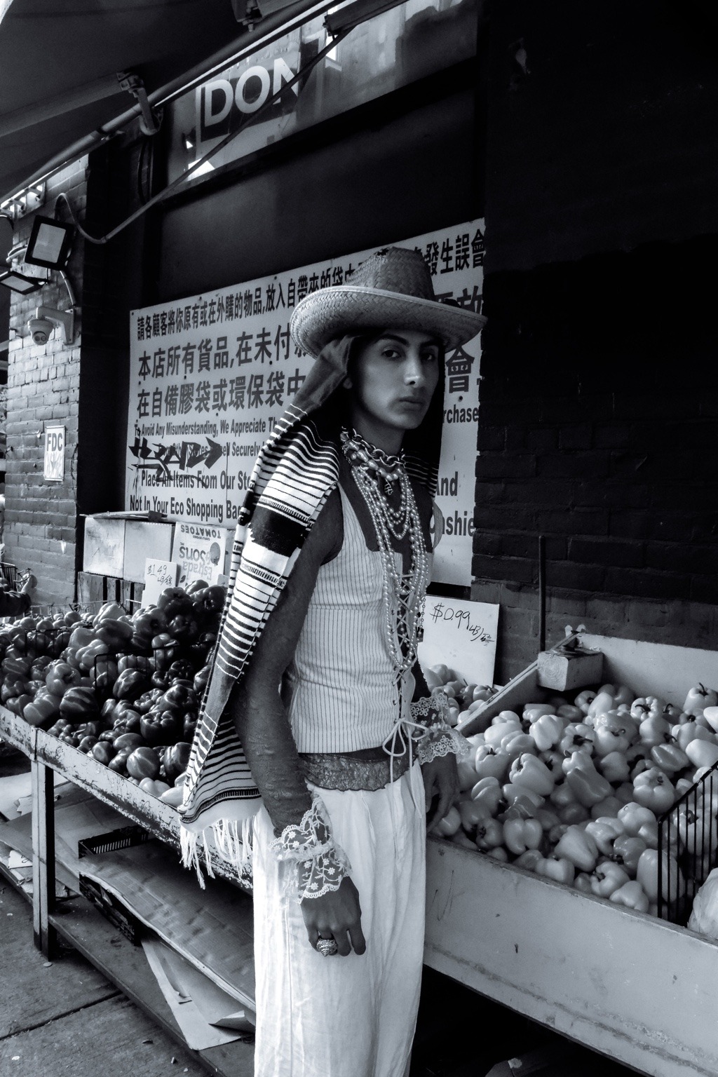 Retrato en blanco y negro con sombrero y joyería frente a un puesto de frutas.