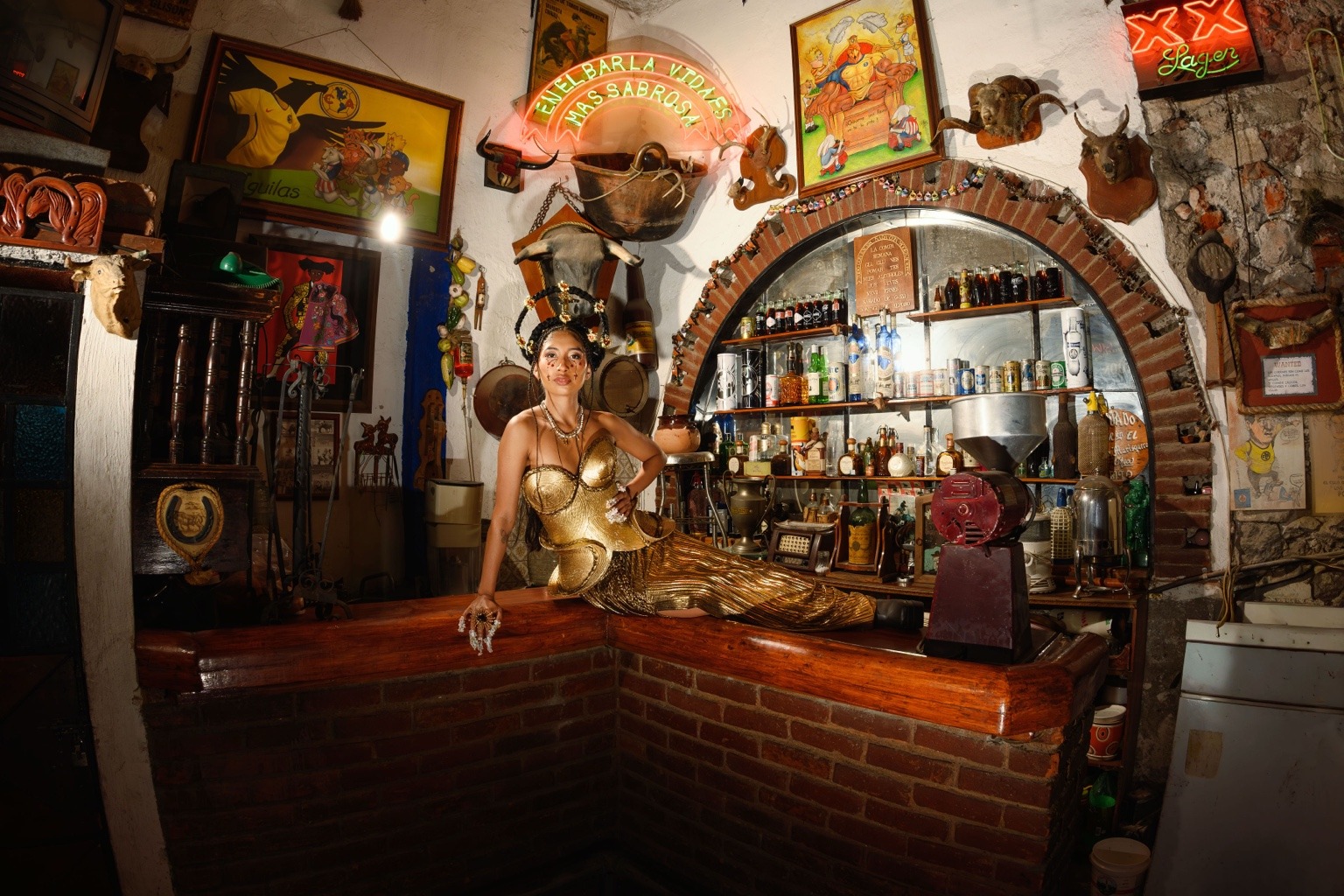 Modelo con look dorado posa sobre la barra rodeada de botellas y objetos.